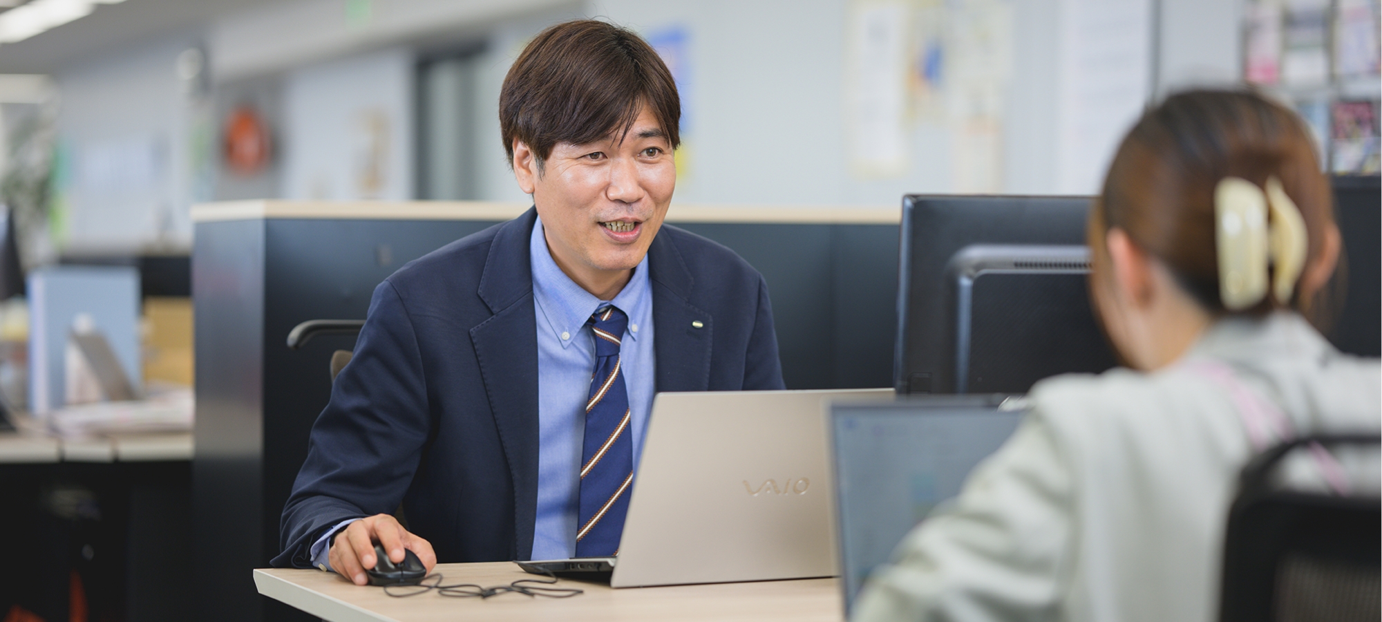 スーツ姿の伊藤 崇がオフィスでコーヒーを片手に、笑顔で同僚と談笑している様子
