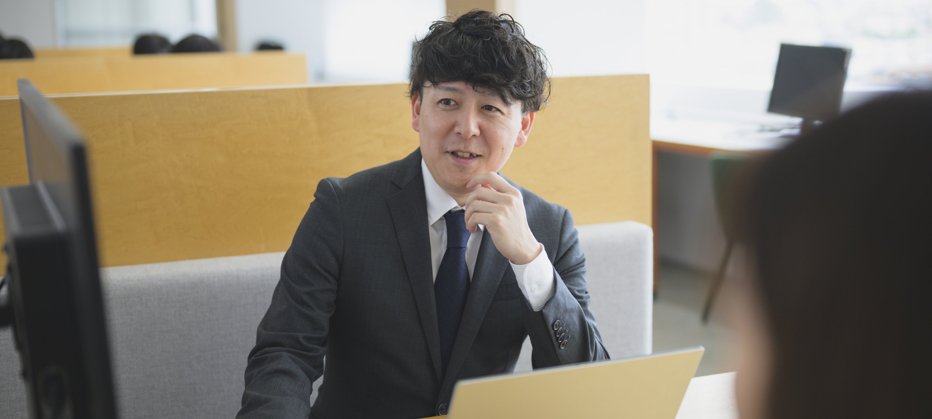 オフィスでパソコンを開き、笑顔で対話するスーツ姿の加藤 雄一