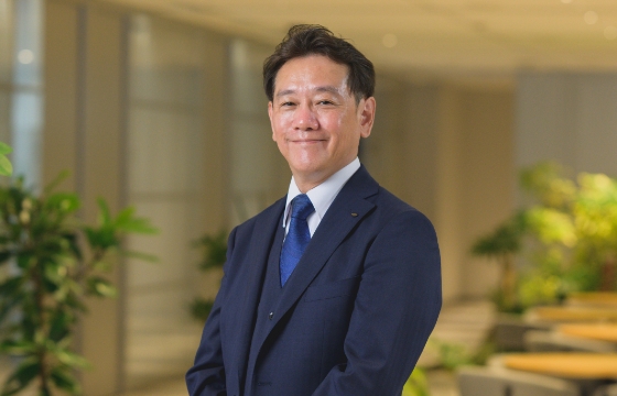 スーツを着た人事総務部長がオフィスの観葉植物の前で微笑んで立っている