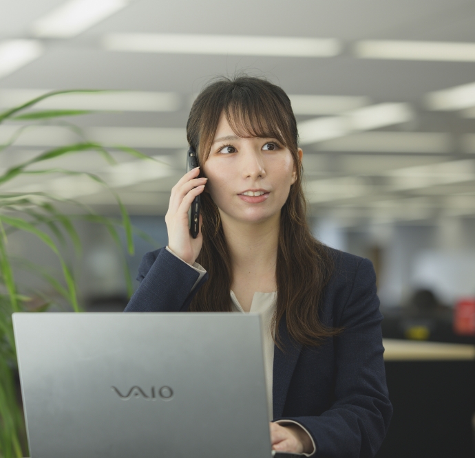 藤澤由佳がデスクでノートパソコンを開きスマートフォンで携帯通話をするスーツ姿