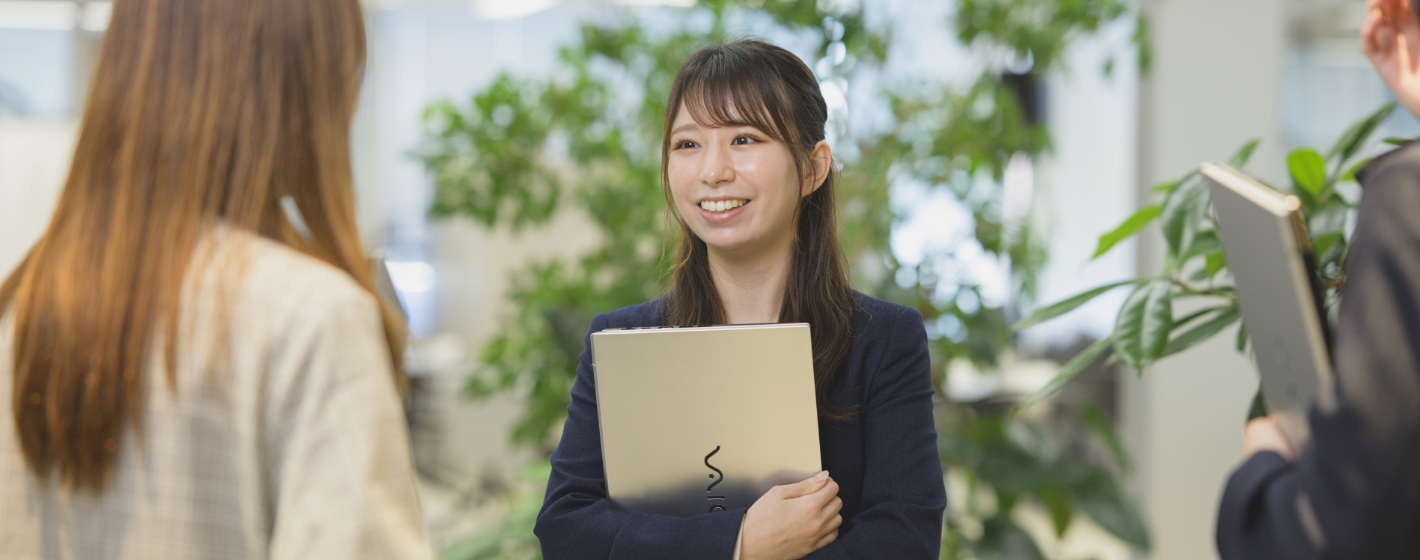 ノートパソコンを抱えて同僚と笑顔で会話するスーツ姿の藤澤由佳