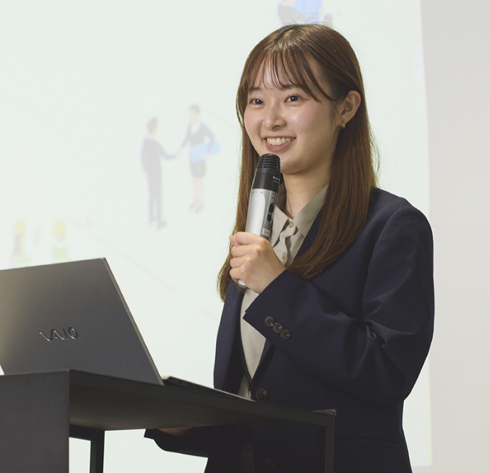 市村春奈がマイクを持ち演台の前で笑顔でプレゼンをするスーツ姿