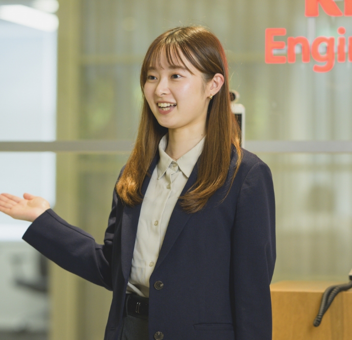手を広げて笑顔で案内をするスーツ姿の市村春奈