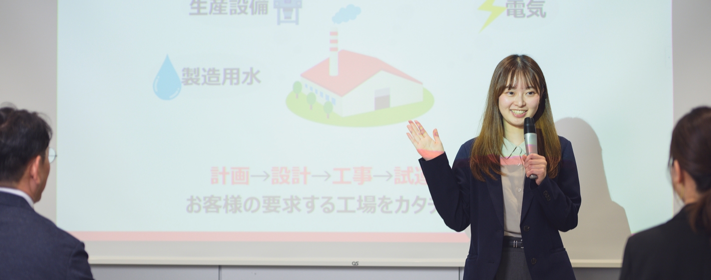 スクリーン前でマイクを持ち笑顔で説明するスーツ姿の市村春奈