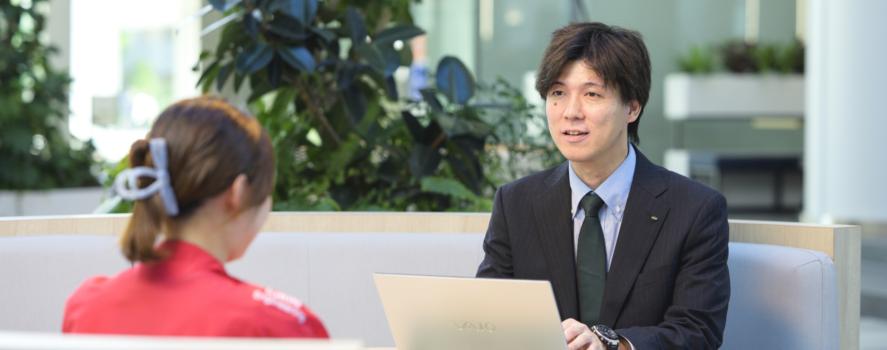 オフィスで同僚と向き合いパソコンを開いて会話をするスーツ姿の甲斐正和