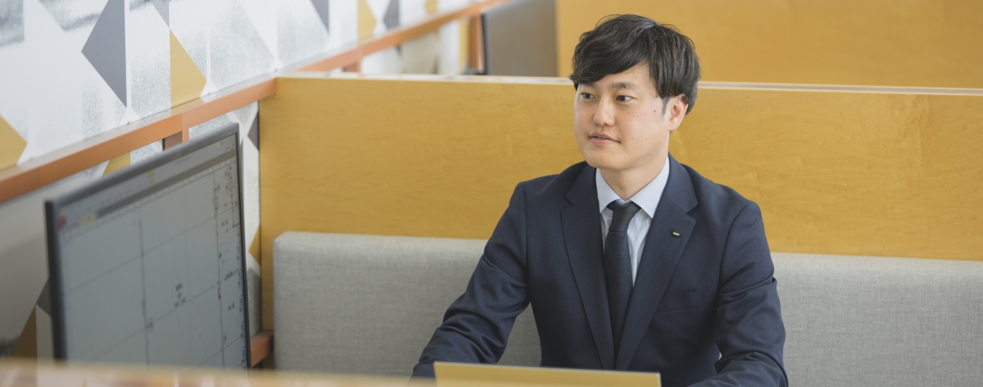 デスクでパソコンに向かい真剣な表情で見つめるスーツ姿の西沢和也