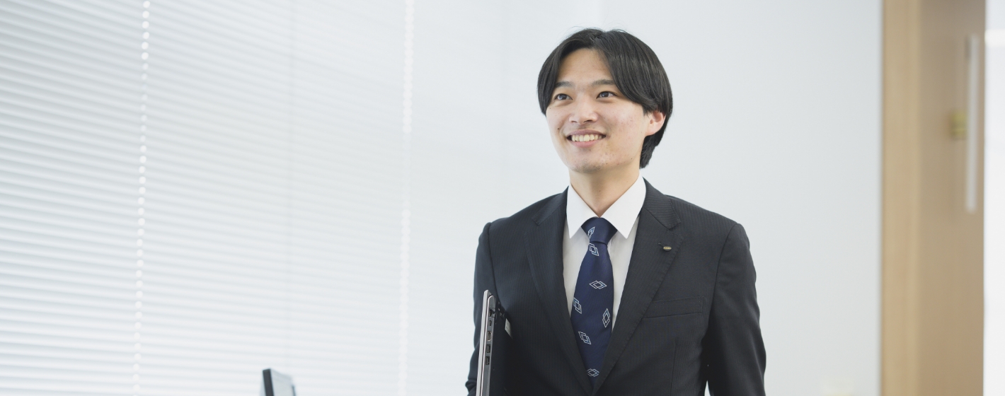 ノートパソコンを持ち笑顔でオフィスを歩くスーツ姿の櫻井敬太
