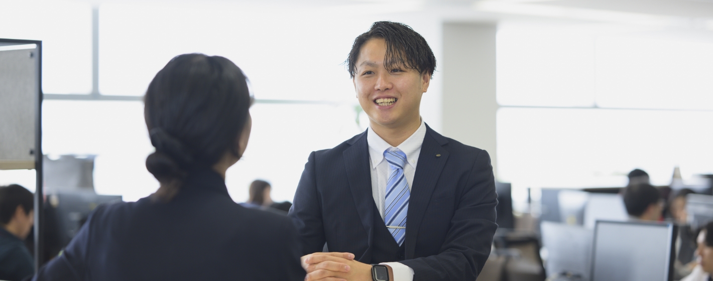 オフィスで同僚と向き合い笑顔で打ち合わせをするスーツ姿の造田 祐斗